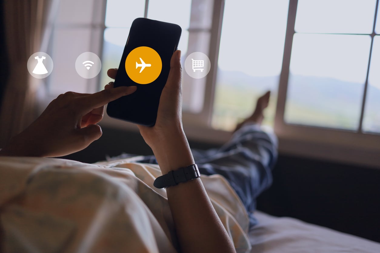 A person lies on a bed using a smartphone with an airplane icon on the screen, surrounded by icons for dress, wifi, and shopping, with large windows in the background.