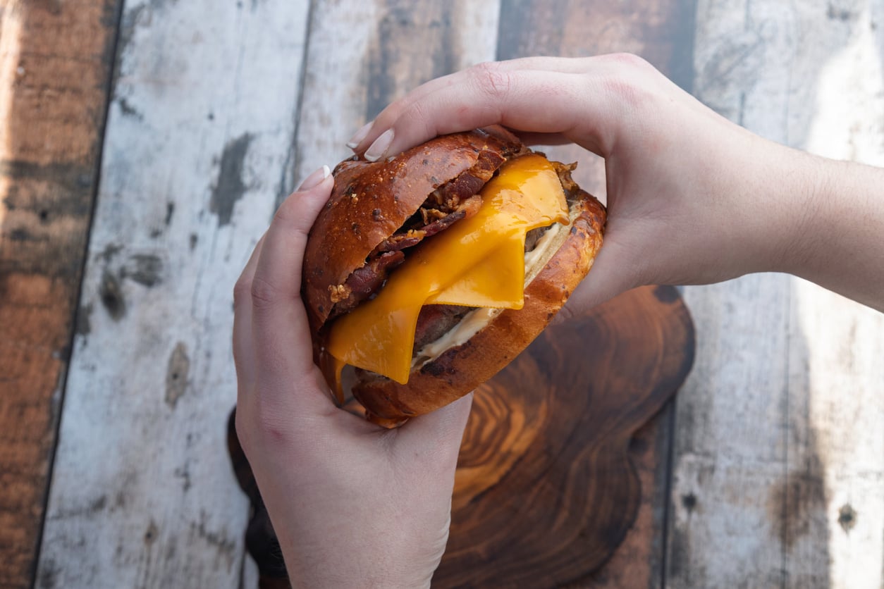 Two hands holding a mouthwatering sandwich, packed with melted cheddar cheese, bacon, and meat inside a glossy bun, over a rustic wooden surface—a true celebration of comfort foods.