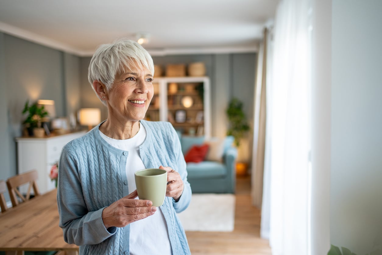 Older woman with short gray hair stands in a cozy, sunlit living room, smiling and holding a mug. Bookshelves, plants, and a sofa are visible in the background, creating a warm, inviting atmosphere.