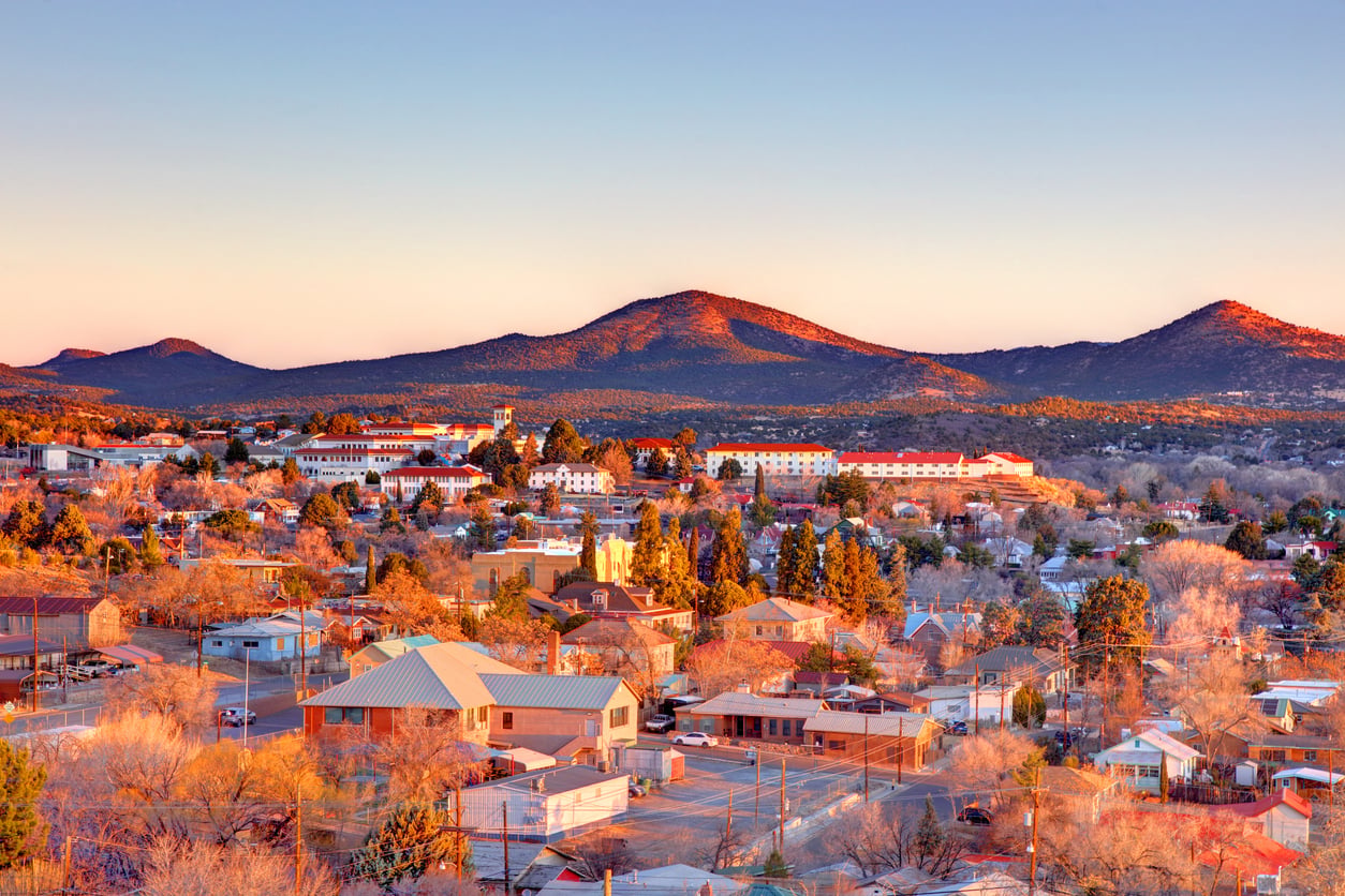 A small town with houses and trees is set against rolling hills and mountains at sunset, with warm sunlight casting a golden glow across the landscape.