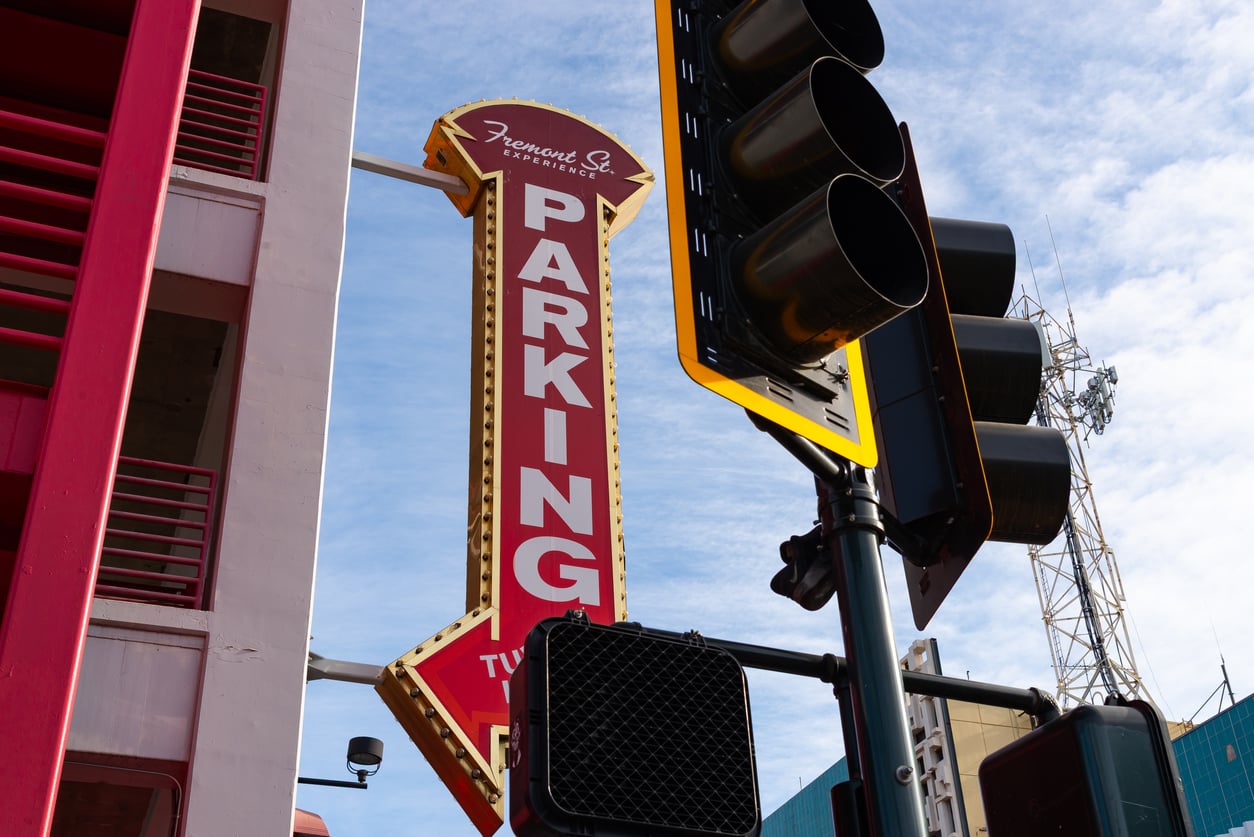 A large red “PARKING” sign with an arrow and a “Fremont St. Experience” label is next to a traffic light and crosswalk signal against a blue sky.