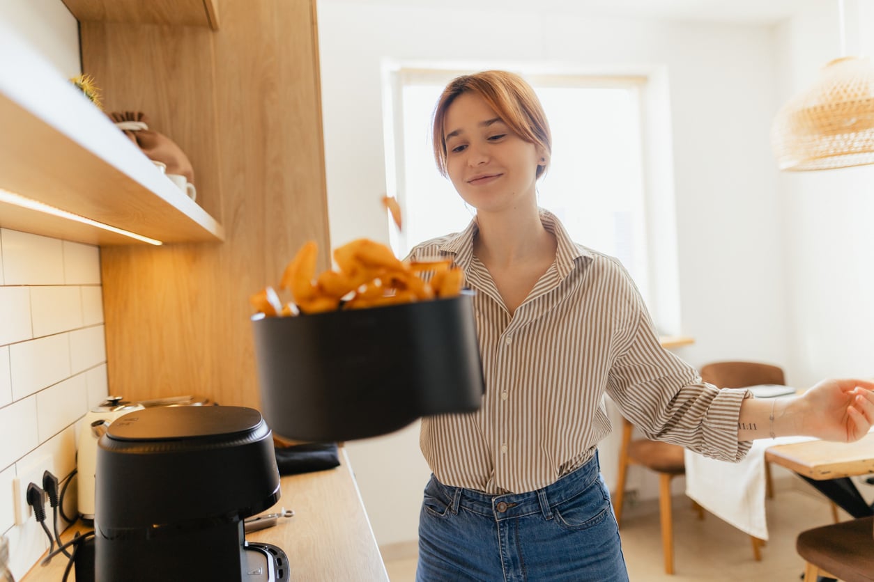 A woman in a striped shirt smiles while holding a basket of freshly cooked fries above an air fryer in a bright kitchen.