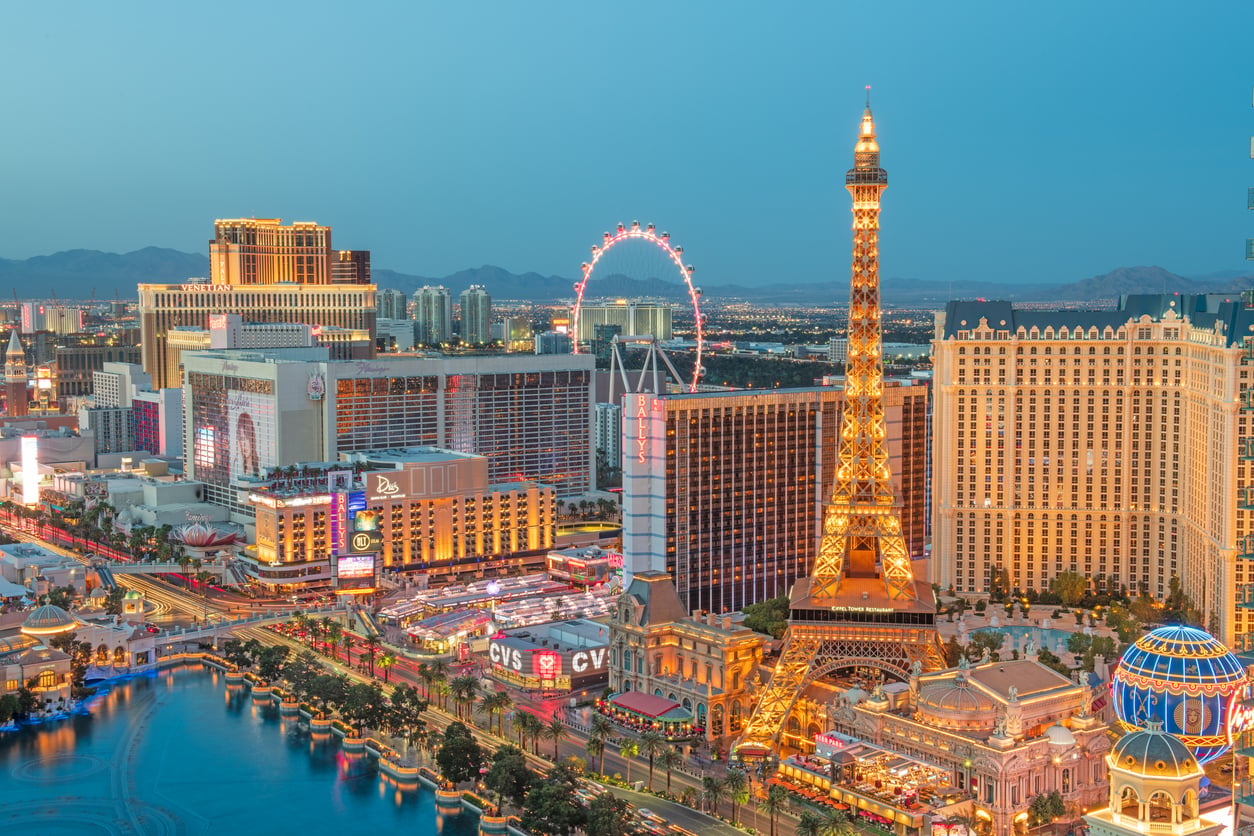 Las Vegas Strip at dusk with bright lights, the Eiffel Tower replica, tall hotels, casinos, and the High Roller Ferris wheel in the background.