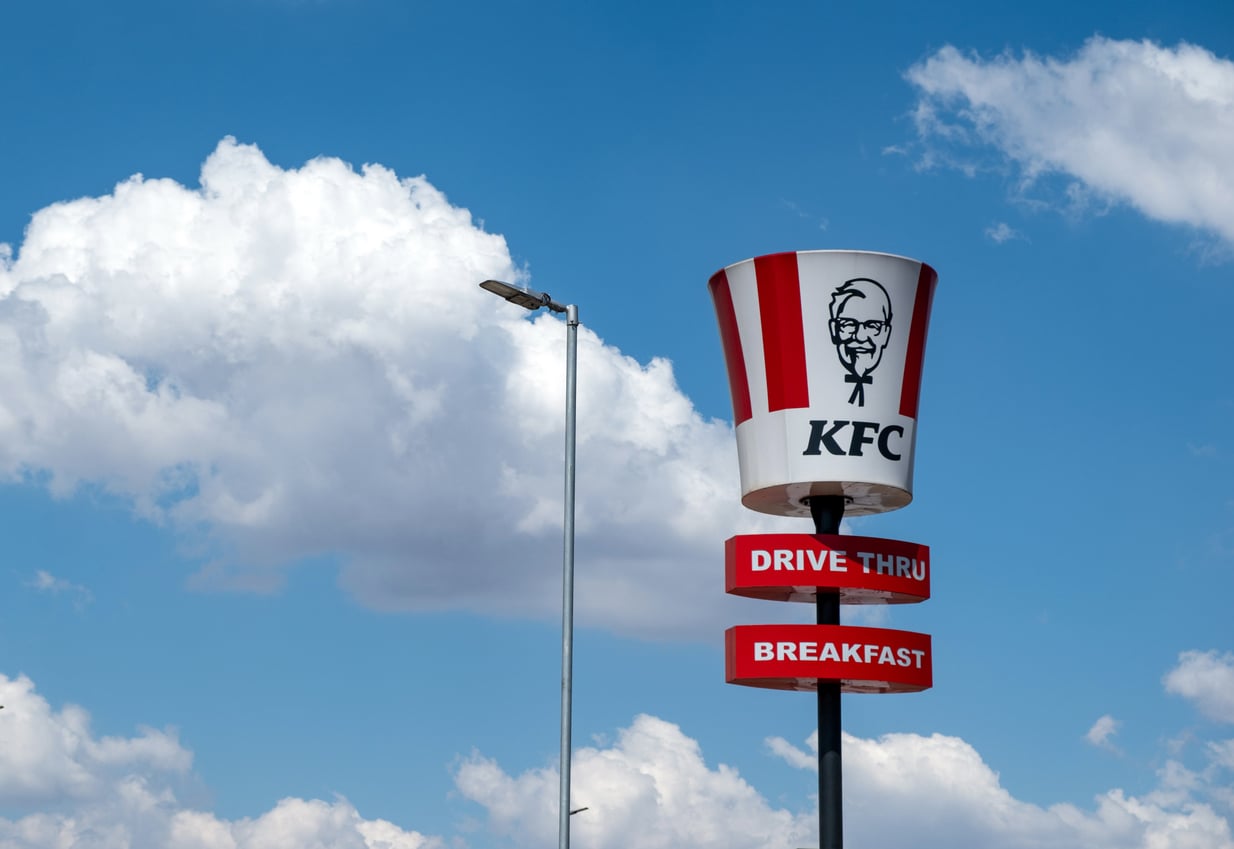 A large KFC bucket sign with “Drive Thru” and “Breakfast” below it stands against a blue sky with scattered clouds.