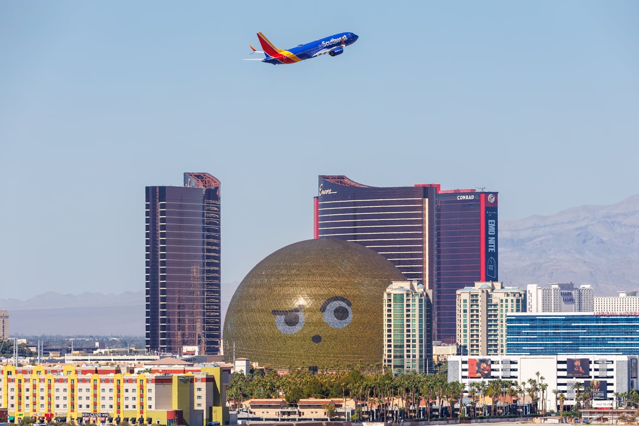A Southwest airplane flies over the Las Vegas skyline, featuring the golden MSG Sphere displaying a winking face, with tall hotels and mountains in the background.