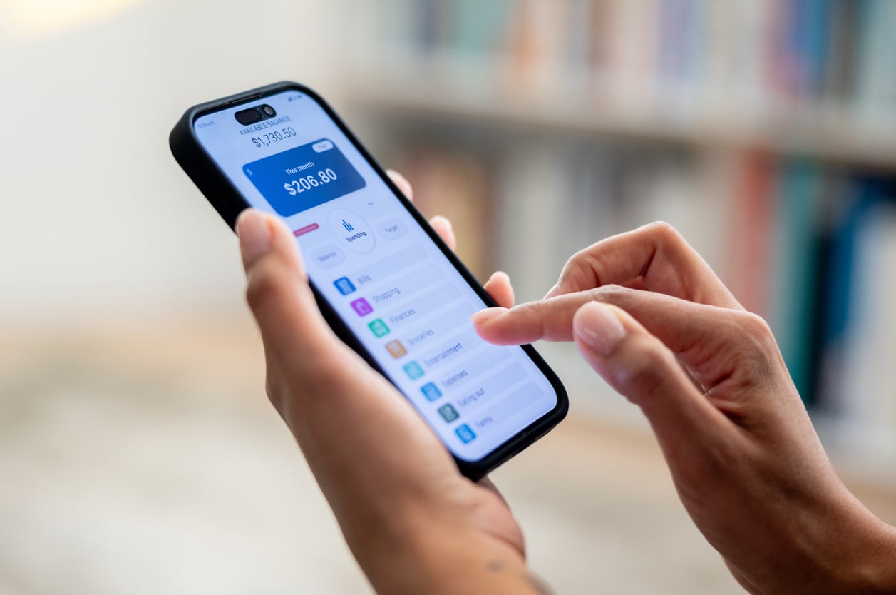 A person uses a smartphone to manage finances, with a banking app displaying account balance and transaction options on the screen.
