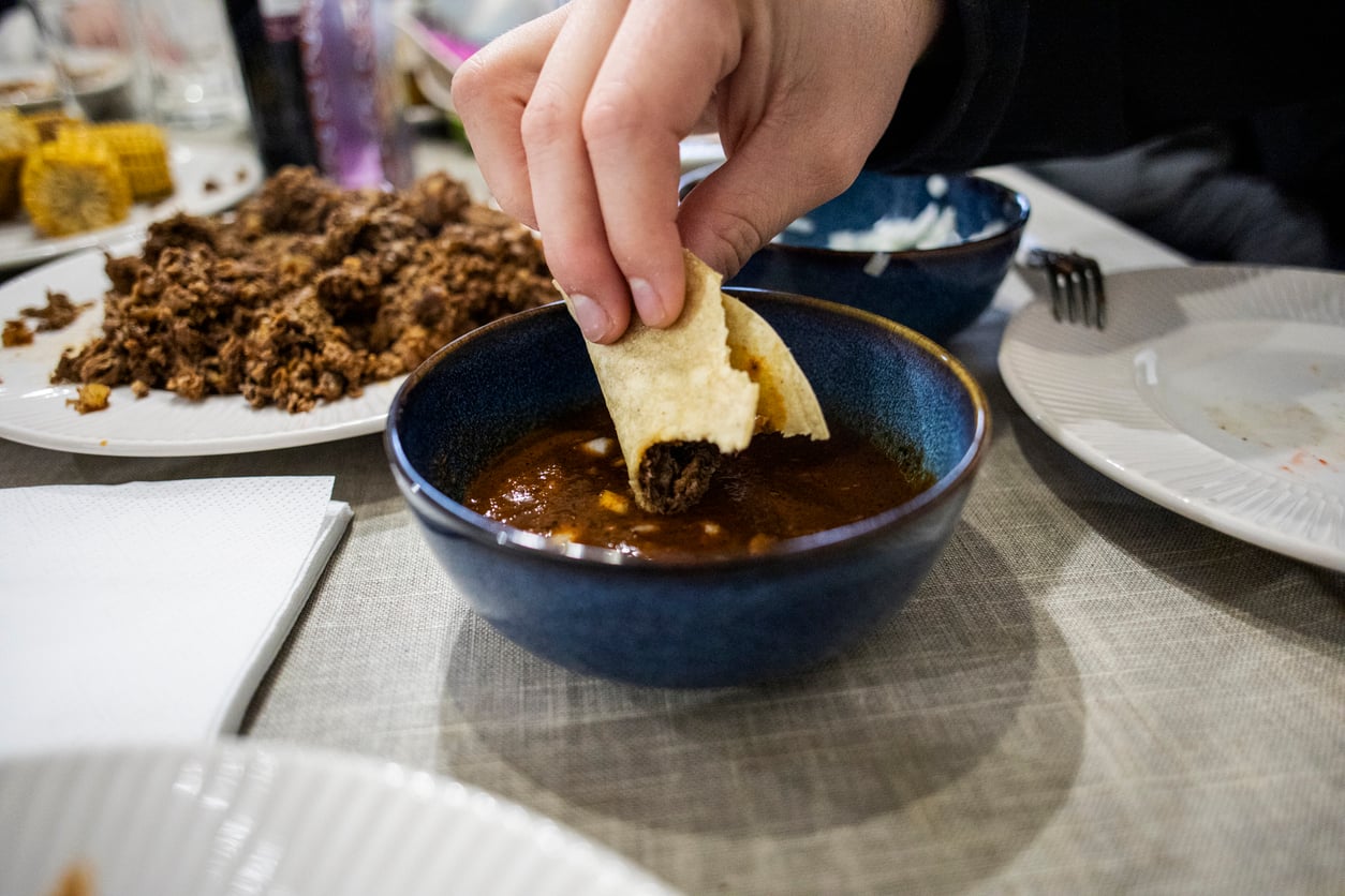 A hand dips a taco filled with shredded meat into a bowl of reddish-brown sauce—one of the must-try foods to order at this spot. Plates with food, napkins, and utensils are visible on the table in the background.