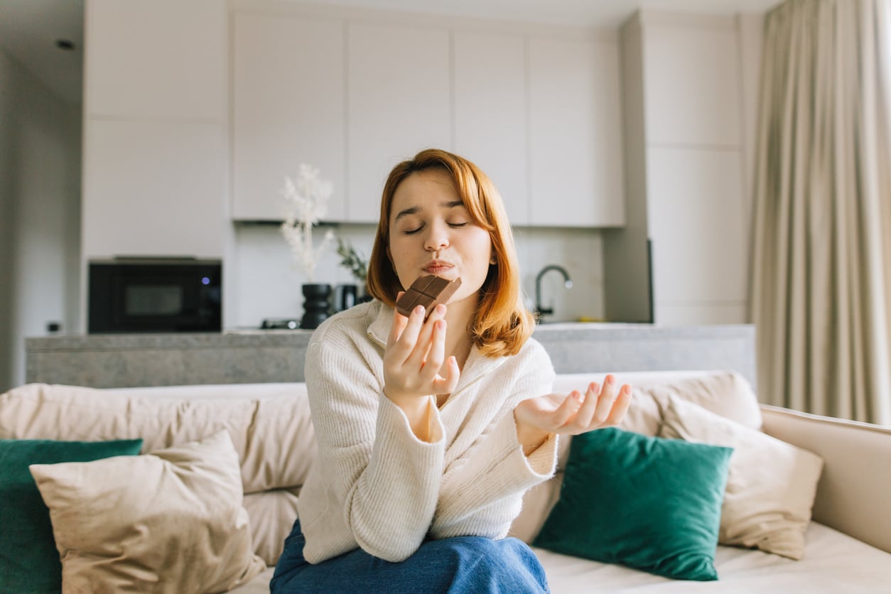 A woman with light brown hair sits on a couch in a modern living room, holding and smelling a piece of chocolate with her eyes closed, appearing to enjoy its aroma.
