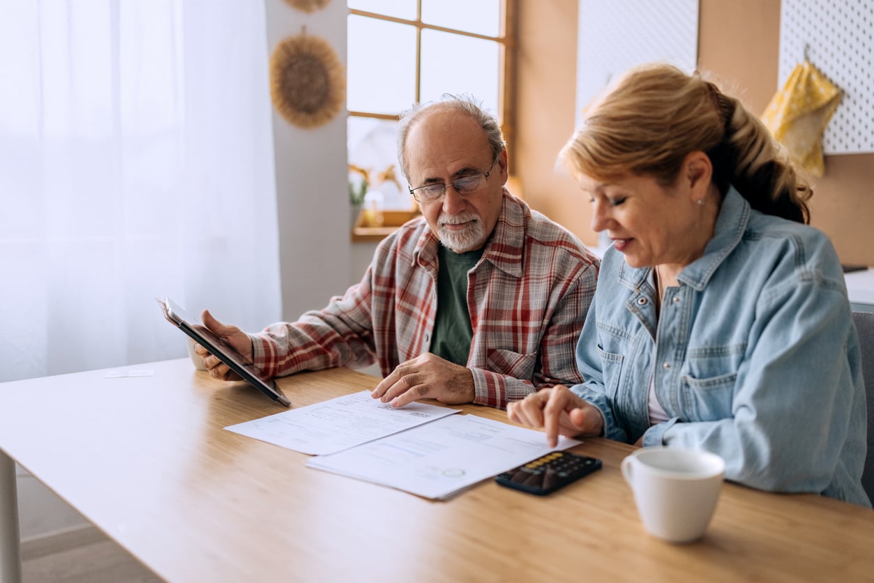 An older man and woman sit at a table reviewing documents. The woman uses a calculator, while the man holds a tablet. Sunlight streams through a window behind them.