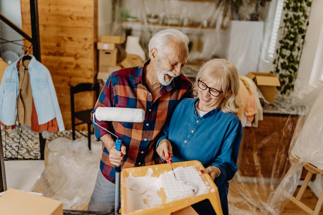 An elderly couple of Baby boomers skills smiles while painting a room. The man holds a paint roller, and the woman holds a paint tray. Moving boxes and covered furniture are visible in the background.