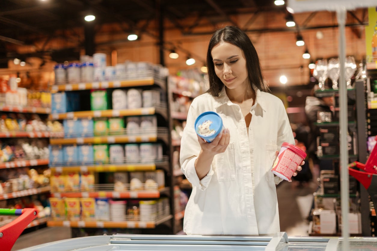A woman in a white shirt stands in a grocery store aisle, holding a blue container in one hand and a pink container in the other, reading the label on the blue container. Shelves of products are in the background.
