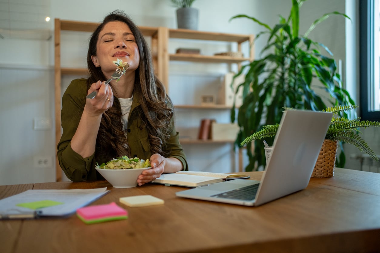 A woman sits at a desk with a laptop, enjoying a bowl of salad. She looks content and relaxed, surrounded by papers, notebooks, and a potted plant in a bright, modern workspace.