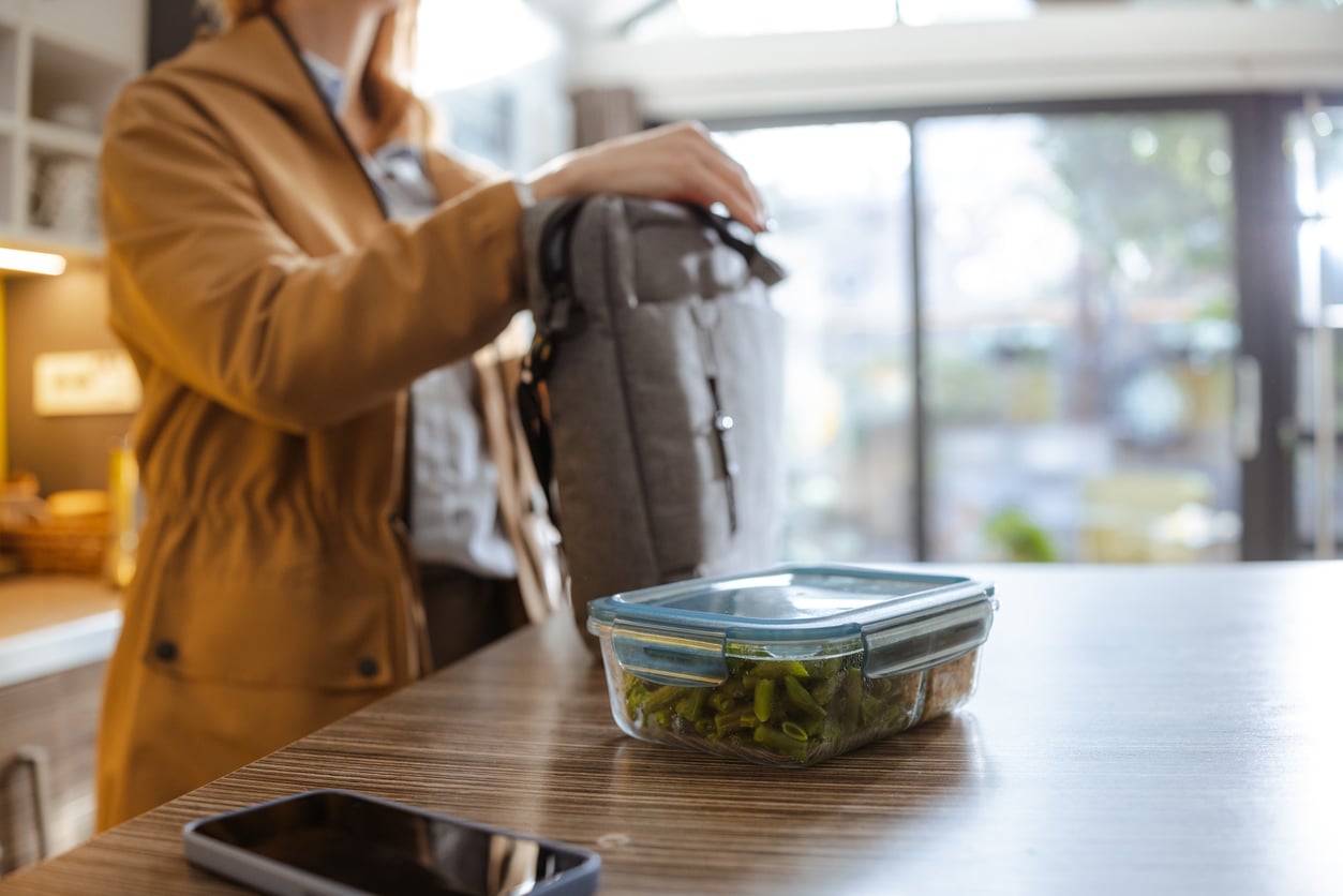 A person in a brown coat stands indoors, placing a gray bag on a table with a container of green beans and a smartphone. Sunlight streams through large windows in the background.