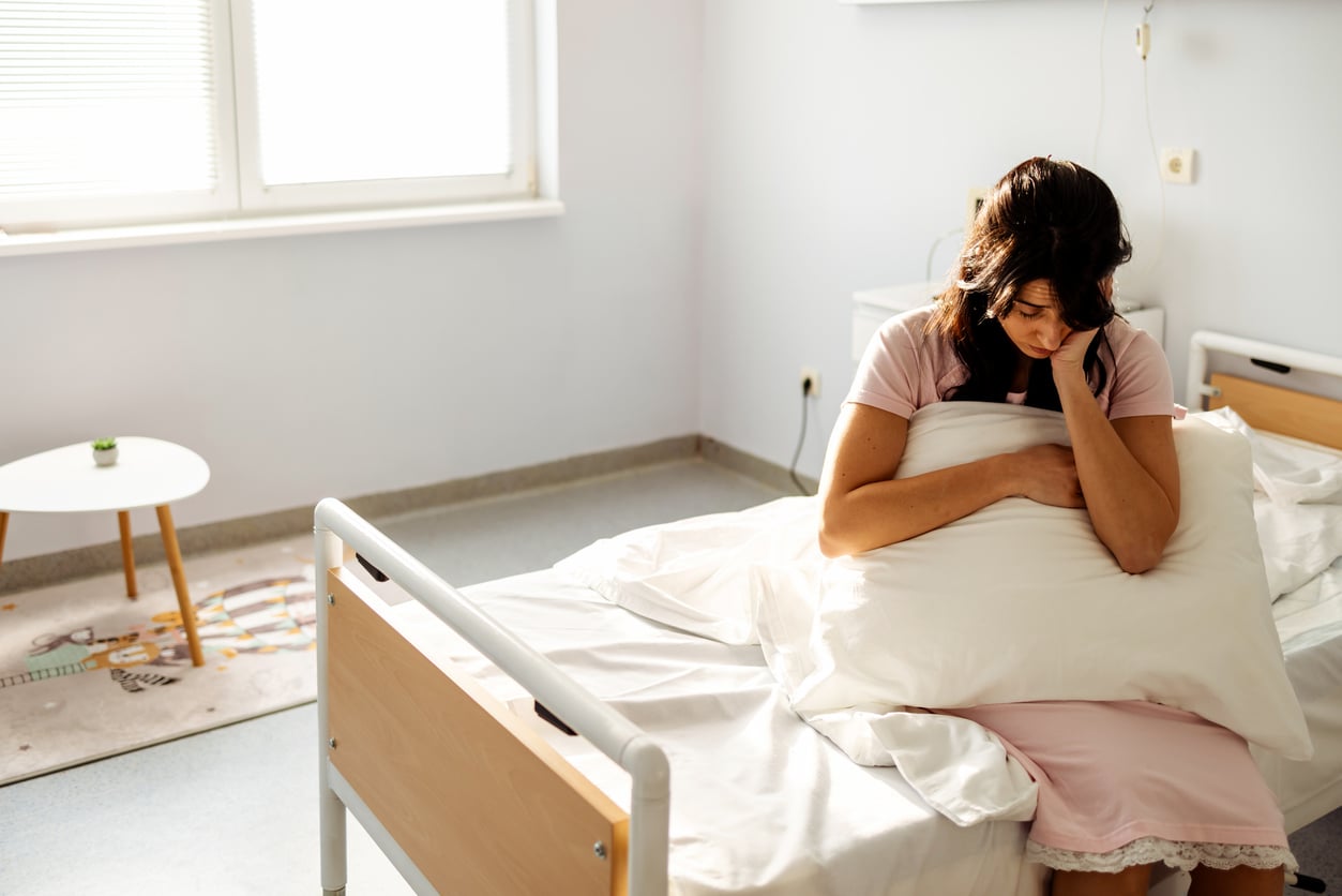 A woman in a pink dress sits on a hospital bed, hugging a pillow and looking down with a sad expression in a bright, sparsely furnished room.