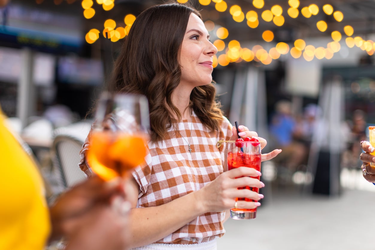 Woman in a gingham top holds a red cocktail with berries and a straw, smiling and talking outdoors with string lights in the background. Other hands holding drinks are visible.