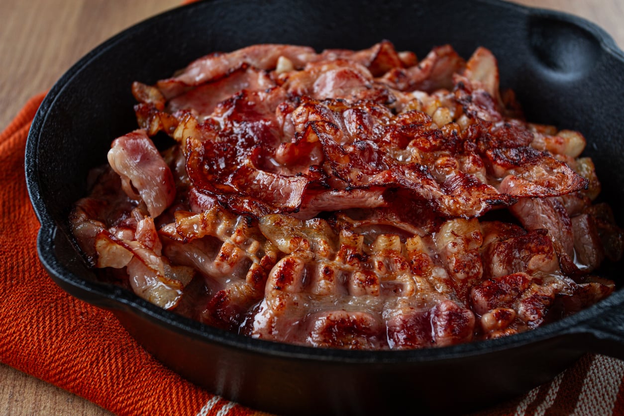 A cast iron skillet filled with crispy, cooked bacon sits on an orange cloth, showcasing one of the most beloved comfort foods, with the bacon glistening and slightly curled, ready to be served.