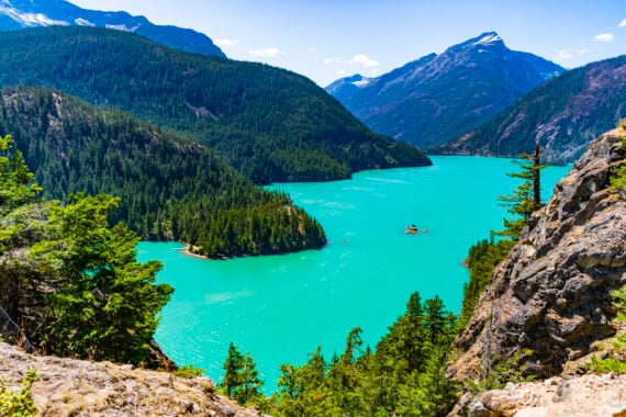 A vibrant turquoise lake surrounded by dense green pine forests and rugged mountains under a bright blue sky with a few clouds.