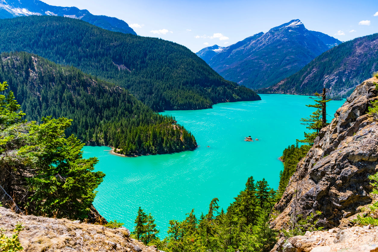 A vibrant turquoise lake surrounded by dense green pine forests and rugged mountains under a bright blue sky with a few clouds.