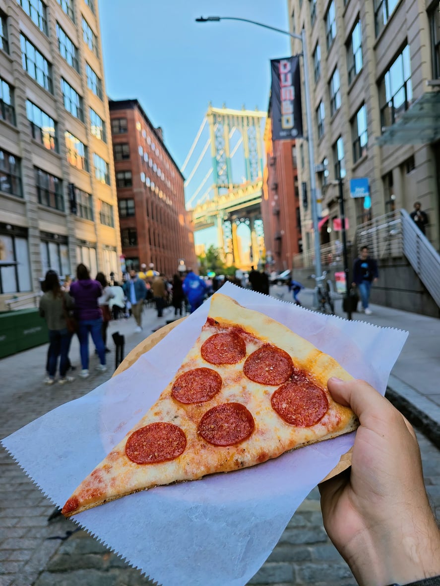 A hand holds a slice of pepperoni pizza on a city street with the Manhattan Bridge visible between tall buildings in the background.