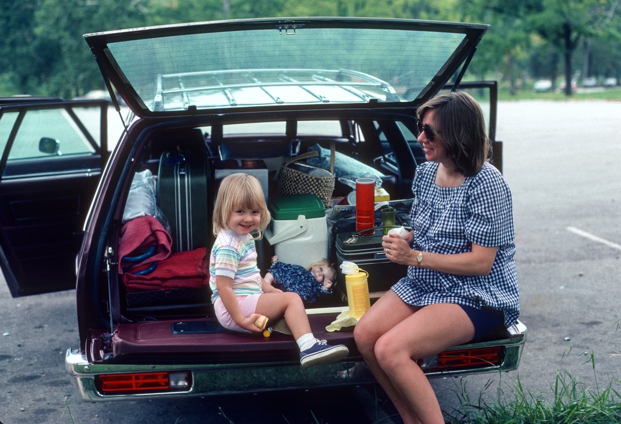 A woman and a young girl sit on the open tailgate of a station wagon filled with picnic supplies, smiling and enjoying a snack in a parking area surrounded by greenery.
