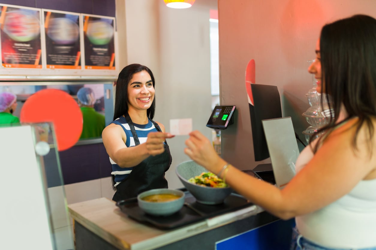 A smiling woman in an apron hands a customer a receipt at a restaurant counter with bowls of food in front of them.