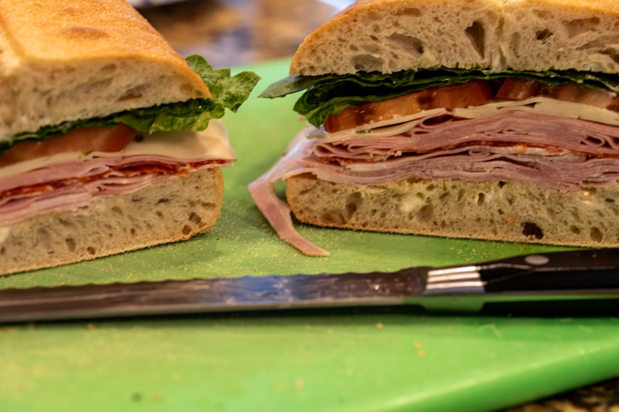 A sandwich with layers of meat, cheese, lettuce, and tomato on ciabatta bread is sliced in half on a green cutting board, with a serrated knife in front.