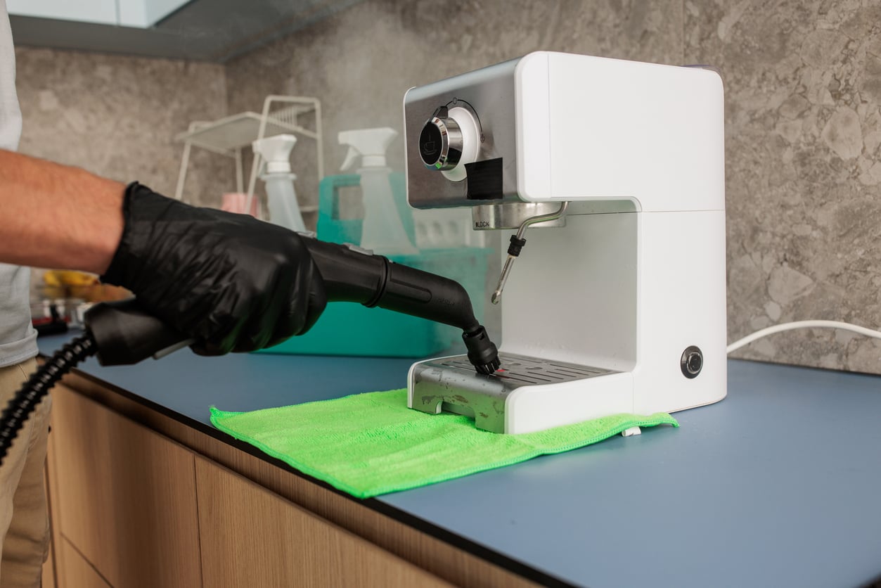 A person wearing a black glove cleans a white espresso machine with a steam cleaner on a green cloth in a modern kitchen.