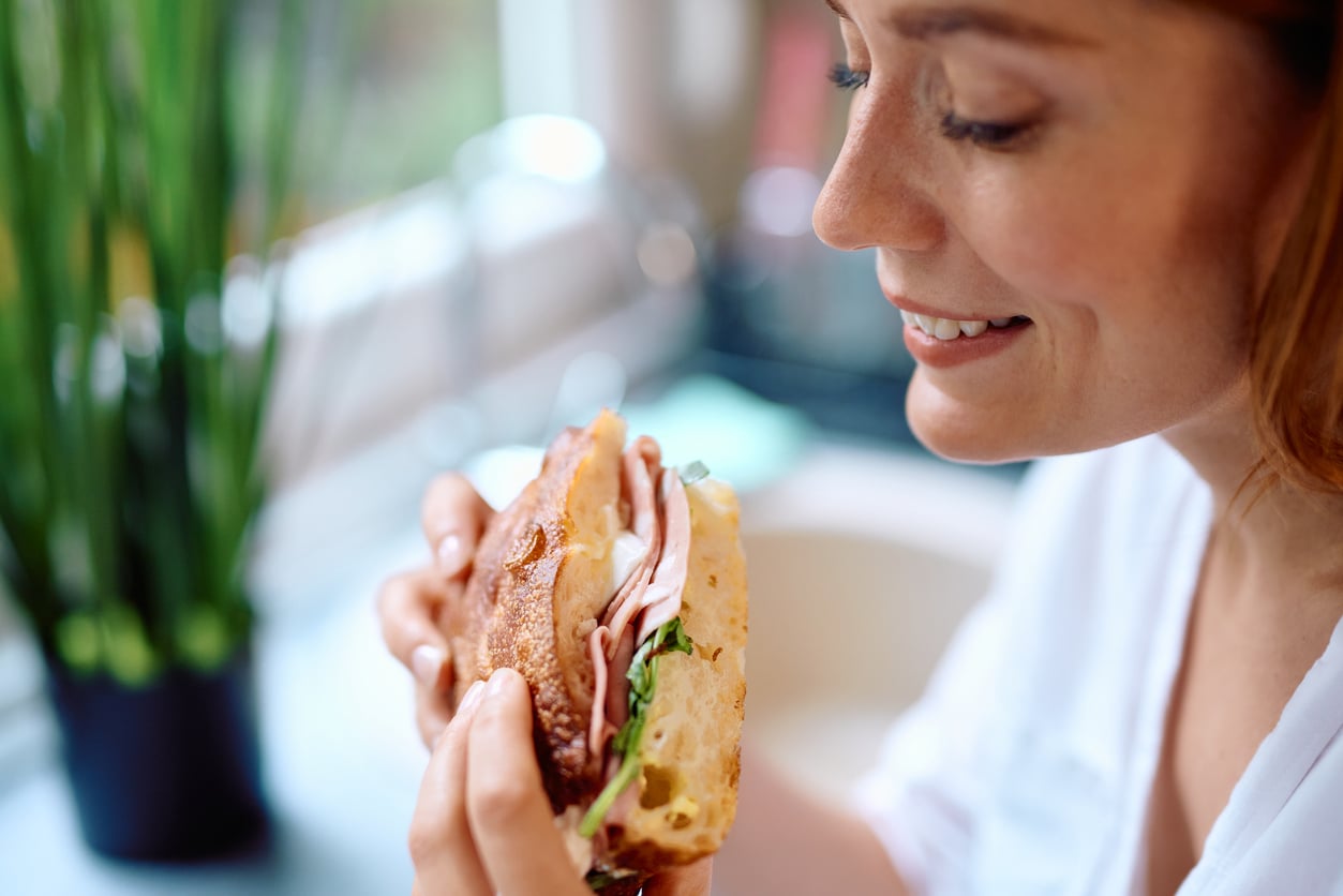 A woman smiles while holding a sandwich filled with deli meat, cheese, and greens. She is indoors, next to a window, with a potted plant blurred in the background.