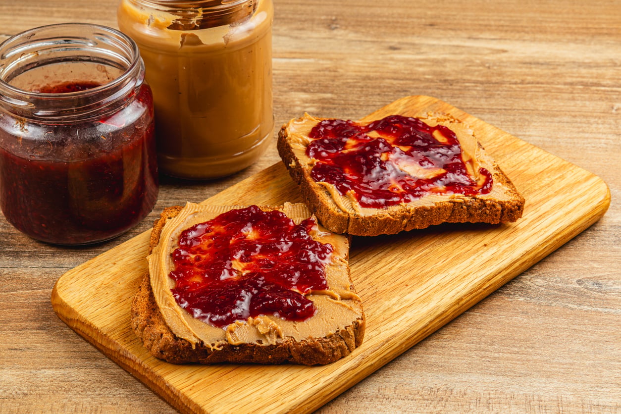 Two slices of bread with peanut butter and jelly on a wooden board, next to open jars of peanut butter and jelly on a wooden table.