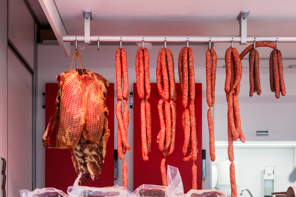 Various sausages and cured meats hang from metal rods in a butcher shop, with a red wall and some equipment visible in the background.