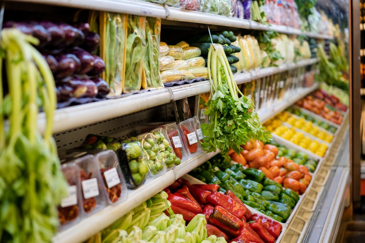 A grocery store produce section displays fresh vegetables—including celery, peppers, tomatoes, cucumbers, and packaged greens—arranged neatly on shelves to inspire healthy grocery habits.