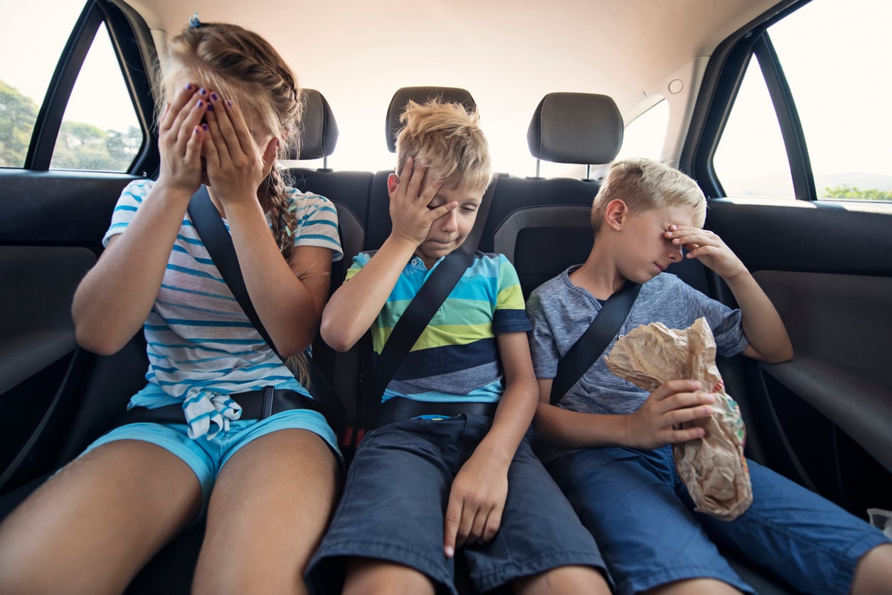 Three children sitting in the back seat of a car during a vacation, all looking unwell with their hands on their faces. One child holds a brown paper bag, possibly feeling carsick. All are wearing seatbelts.