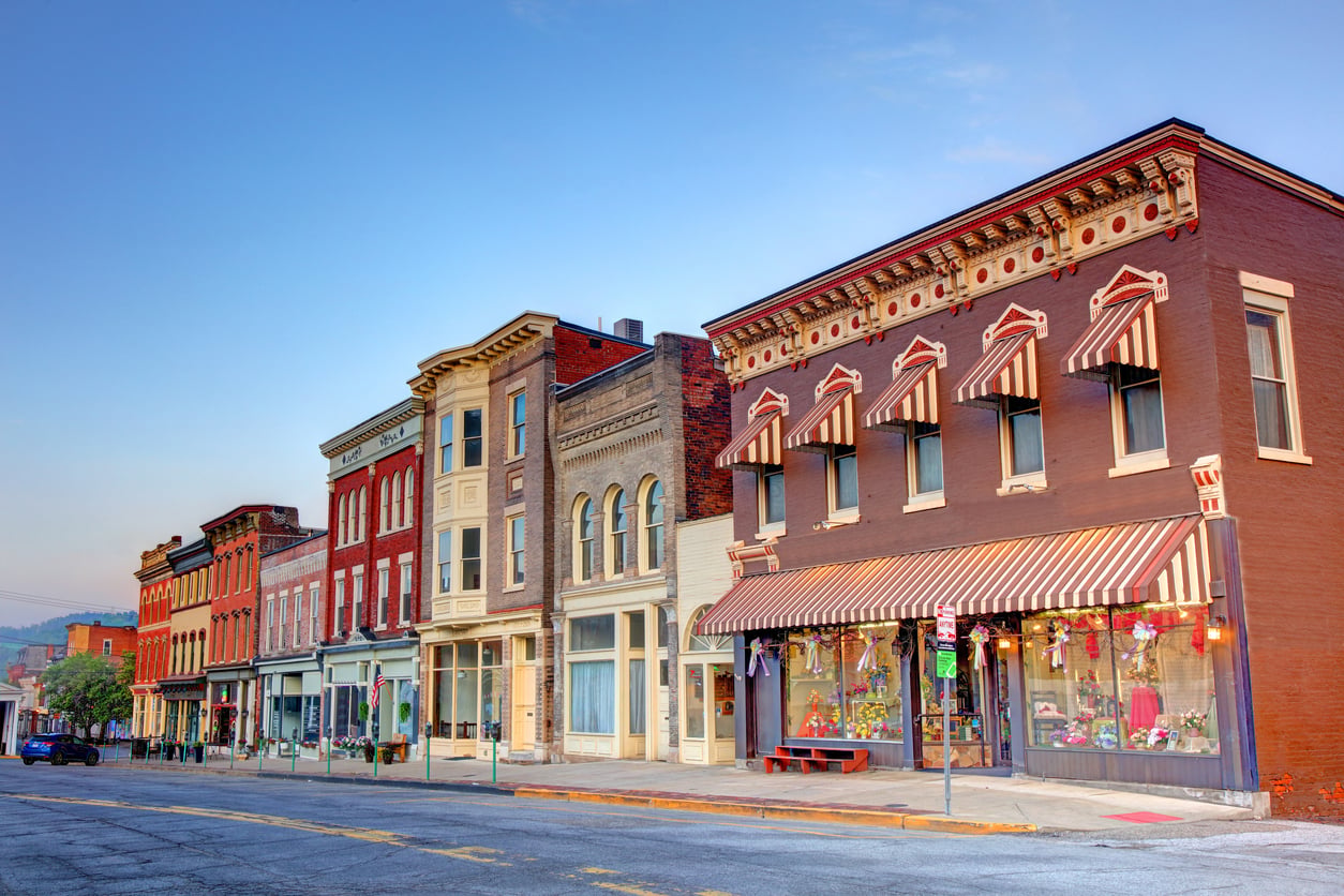Colorful historic storefronts line a quiet small-town street at sunset, with striped awnings, large windows, and decorative brickwork under a clear blue sky.