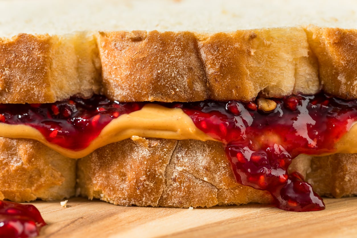 Close-up of a peanut butter and raspberry jelly sandwich on thick, crusty bread, with jelly oozing out the sides onto a wooden surface—a timeless delight among comfort foods.