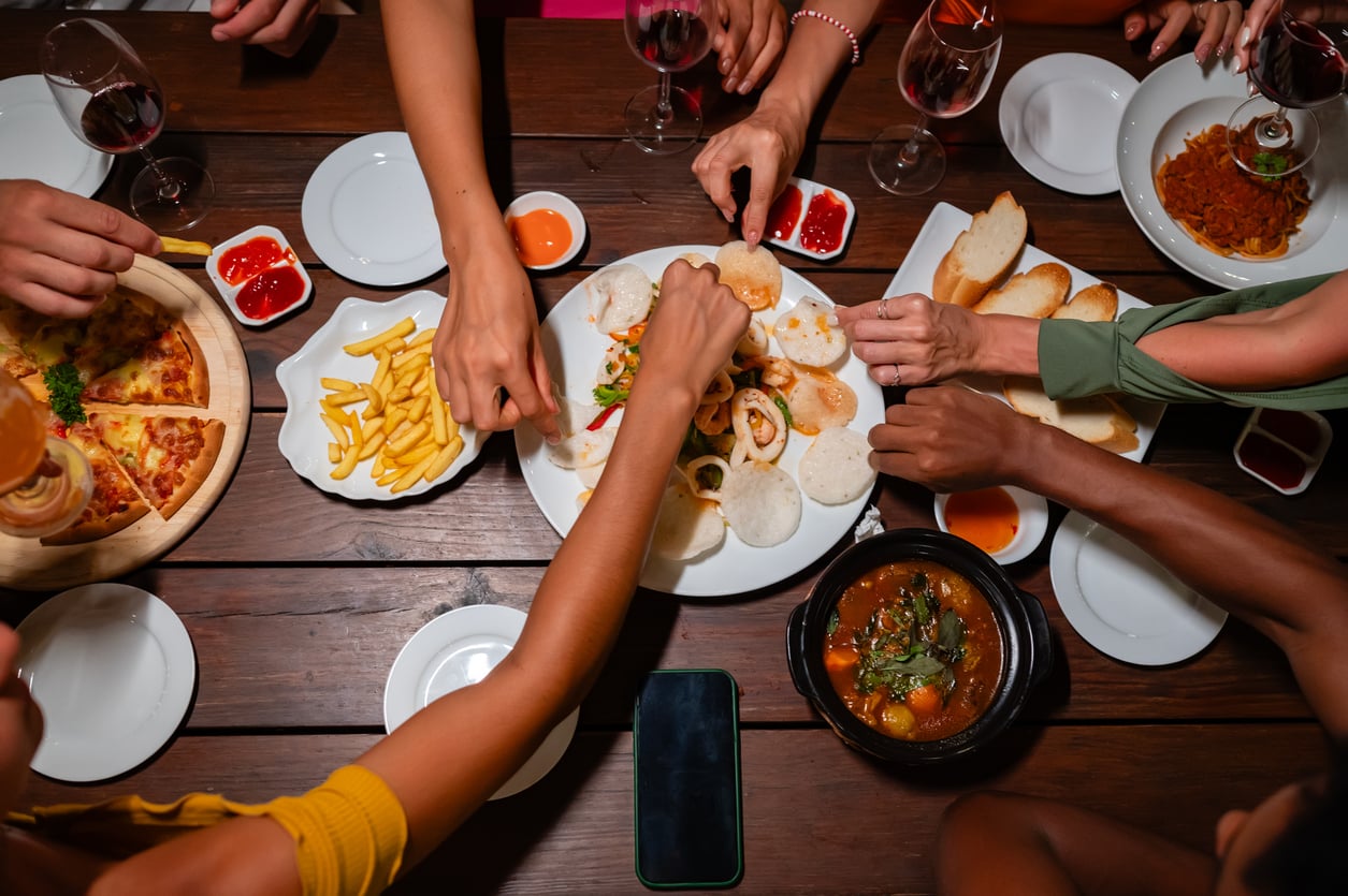 A group of people reach for food on a wooden table topped with pizza, fries, bread, dips, and wine glasses. Hands from different directions gather around shared dishes.