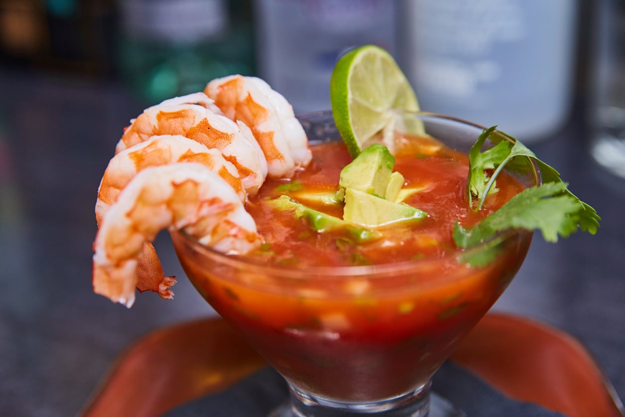 A glass filled with shrimp cocktail, one of the restaurant specialties, is garnished with avocado slices, lime wedges, and cilantro, and sits on a dark surface with blurred bottles in the background.