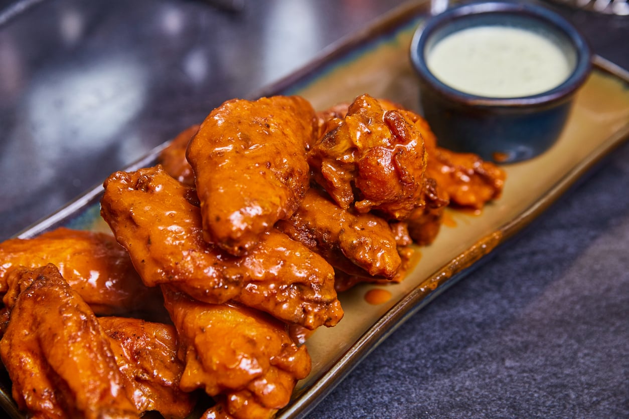 A plate of saucy buffalo chicken wings is served with a small cup of creamy dipping sauce on the side.