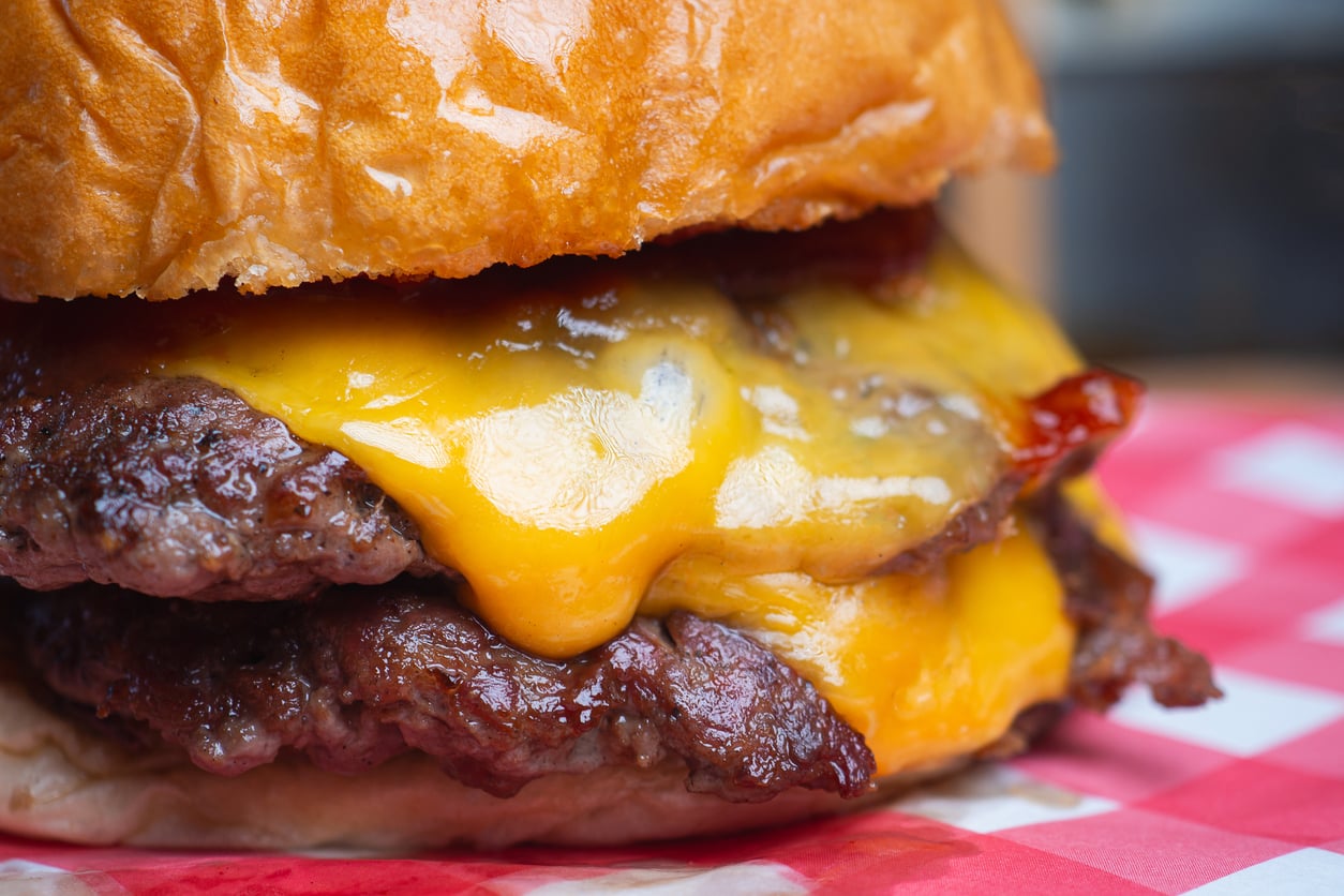 Close-up of a juicy double cheeseburger with melted cheddar cheese on two beef patties, served on a shiny toasted bun, resting on a red and white checkered paper.