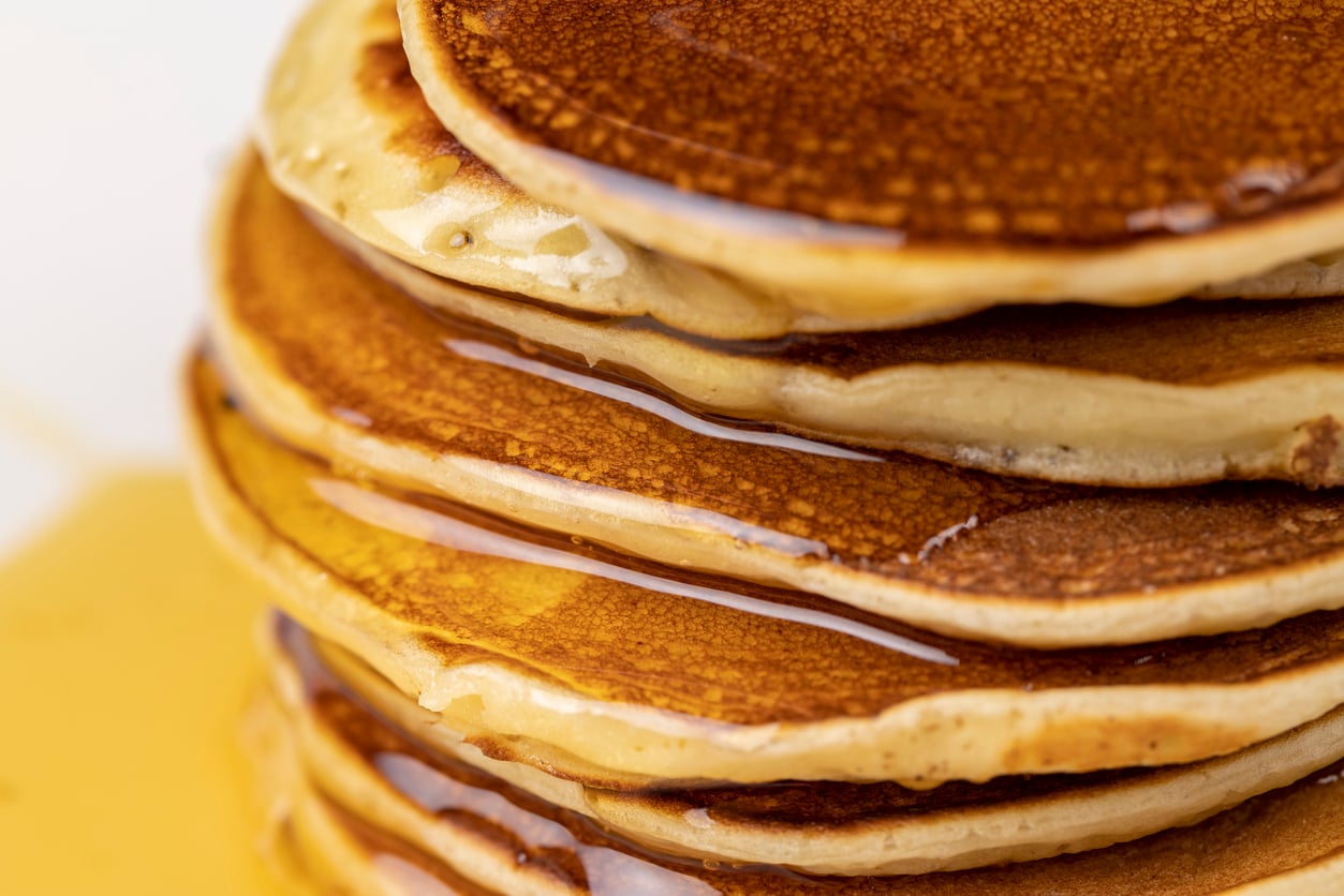 A close-up of a stack of golden brown pancakes covered in syrup, highlighting their fluffy texture and the glistening surface of the syrup.