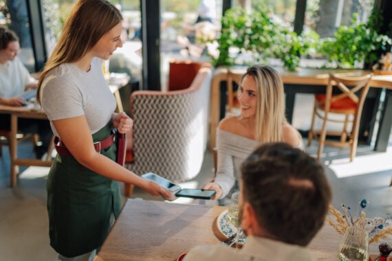 A smiling woman in a restaurant pays with a card using a payment terminal held by a waitress in a green apron. Another person sits at the table. Sunlight streams through large windows.