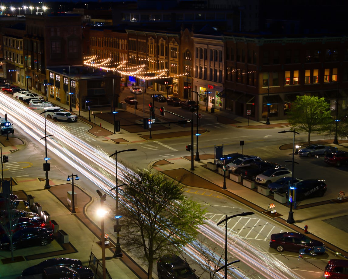 Downtown street at night with light trails from cars, illuminated buildings, string lights, parked cars, and trees along the sidewalk. Urban, lively atmosphere with glowing streetlights and visible crosswalks.