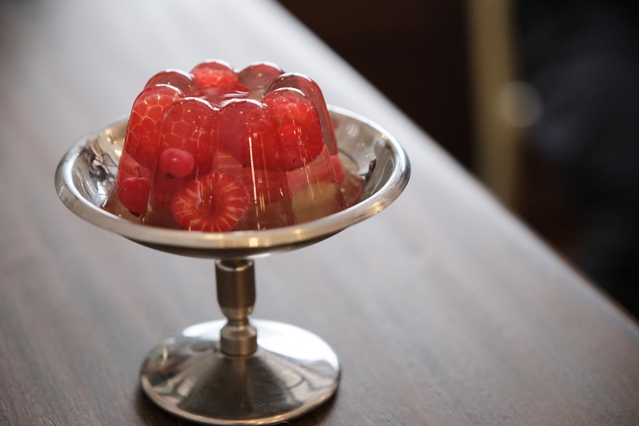 A silver dish holds a clear gelatin dessert with raspberries and red currants set inside, sitting on a wooden surface—a beautiful example of how Everyday Foods That Used to Be Considered Fancy can bring simple elegance to any table.
