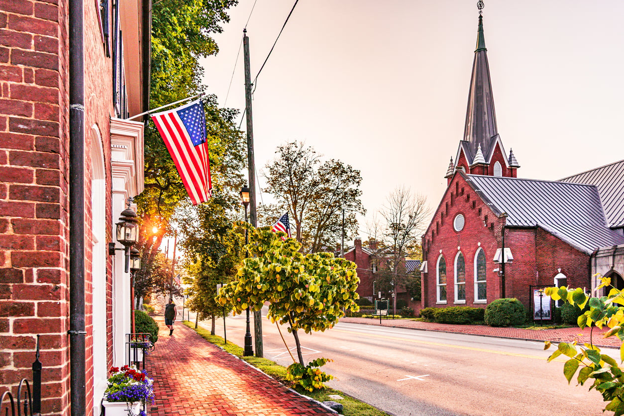A quiet street with American flags, brick buildings, trees, and a church with a tall steeple at sunrise. A person walks on the sidewalk.