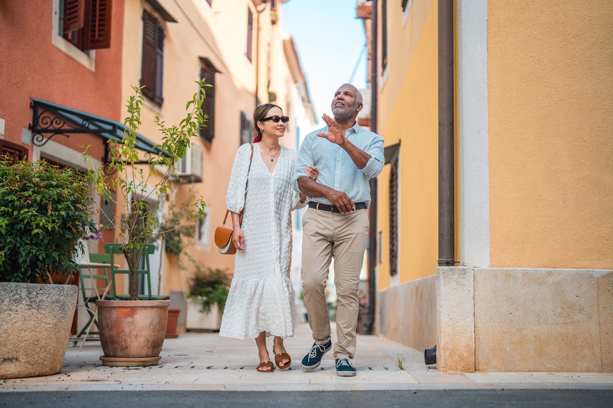 A smiling couple walks arm in arm down a sunny, narrow street lined with colorful buildings and potted plants, savoring unforgettable vacation experiences together.