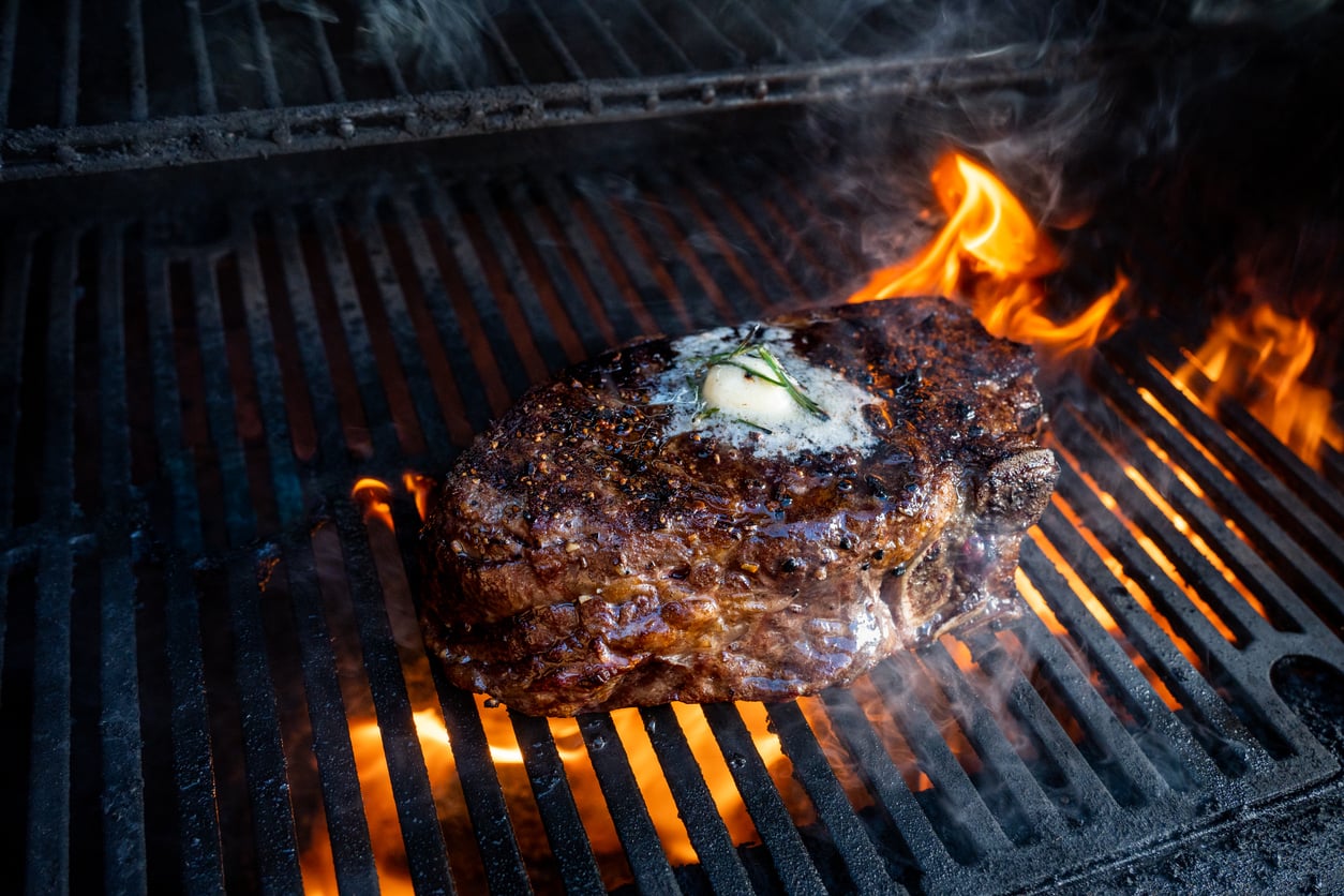A thick steak with melted butter and herbs is grilling over open flames, with char marks and smoke rising from the sizzling meat.