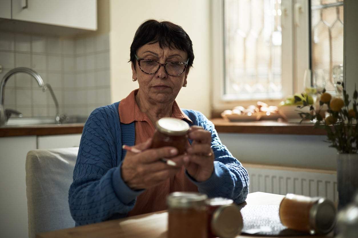 An older woman wearing glasses and a blue cardigan sits at a kitchen table, looking closely at a jar of preserves. Other jars and a notebook are on the table beside her.