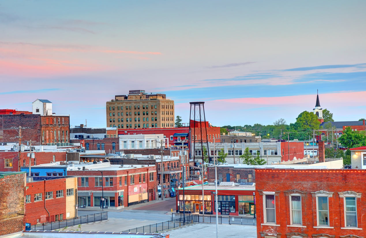 Colorful downtown cityscape at sunset, with brick buildings, a tall structure in the center, and a church steeple visible in the background under a partly cloudy sky.