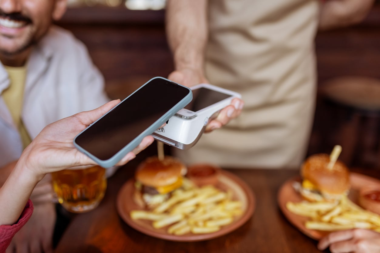A person pays for a meal using a smartphone and a wireless card reader at a restaurant, with burgers and fries on the table.
