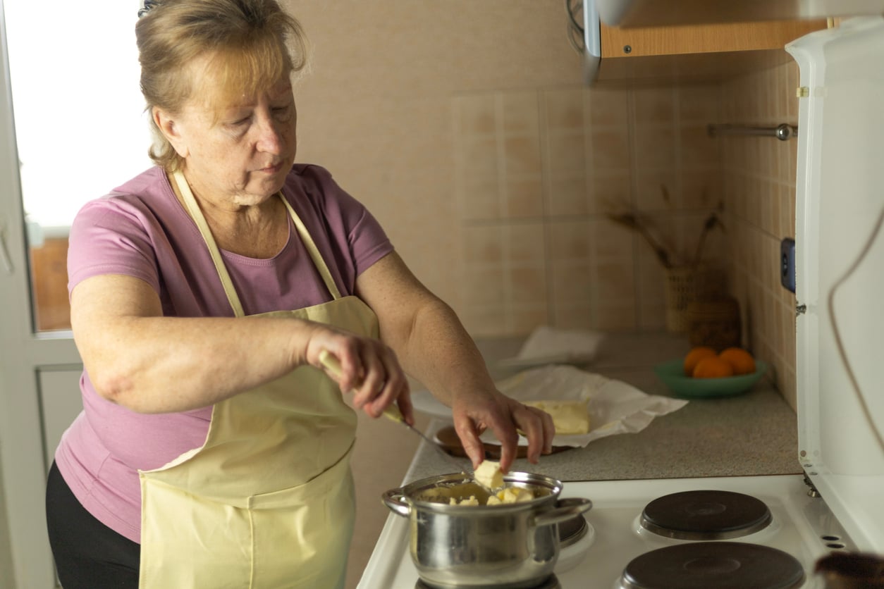 An older woman in a yellow apron and purple shirt is cooking at a stove, placing butter into a pot. The kitchen counter behind her has fruit and some items.