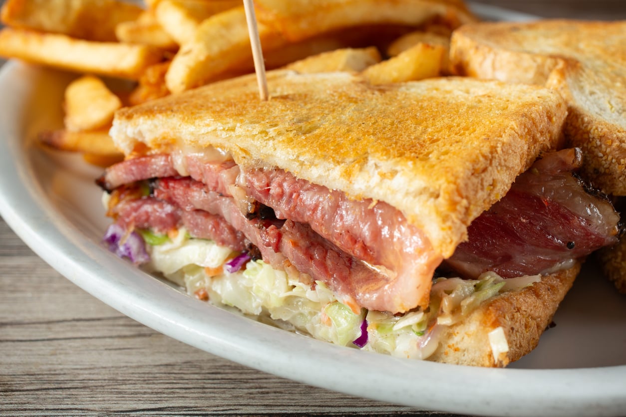 A close-up of a sandwich with sliced meat, coleslaw, and toasted bread on a plate, served with crispy French fries in the background.